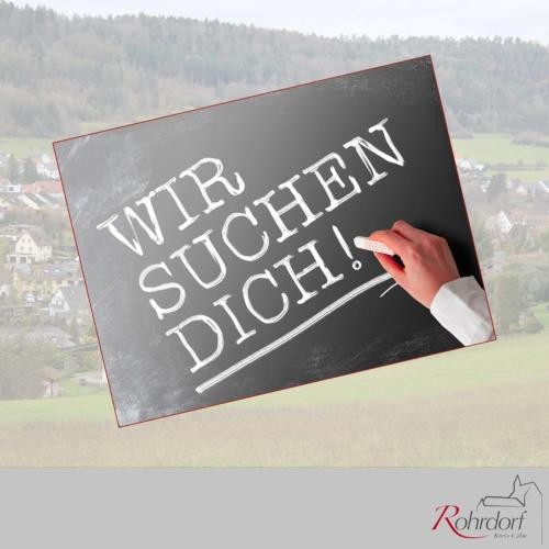 Stellenausschreibung Schwarze Tafel mit der Aufschrift „Wir suchen dich!“ in weißer Kreideschrift, eine Hand schreibt gerade mit Kreide. Im Hintergrund ist unscharf eine Ortsansicht von Rohrdorf zu sehen. Unten rechts befindet sich das Logo der Gemeinde Rohrdorf.