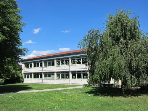 Zweistöckiges Schulgebäude mit langen Fensterreihen, eingebettet in eine Grünfläche mit Rasen und Bäumen. Vor dem Gebäude ein Fußweg, darüber blauer Himmel mit vereinzelten Wolken.