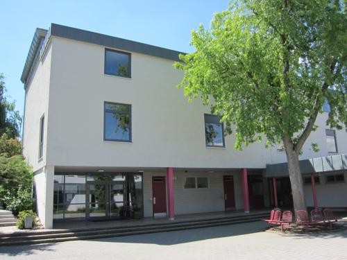 Außenansicht eines modernen Schulgebäudes mit hellem Putz, großen Fenstern und überdachtem Eingangsbereich. Vor dem Gebäude ein gepflasterter Schulhof mit Sitzbänken und einem großen Baum.