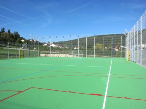 „Mehrzweck-Sportfeld mit grünem Kunststoffbelag und eingezeichneten Linien für verschiedene Sportarten. Das Spielfeld ist von einem hohen Zaun umgeben, im Hintergrund Hügel, Wohnhäuser und blauer Himmel.
