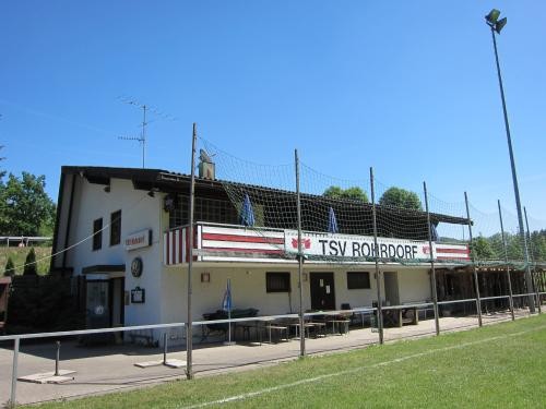 Vereinsheim des TSV Rohrdorf am Sportplatz mit überdachter Terrasse, Tischen und Bänken. An der Fassade ein großes Banner mit der Aufschrift ‚TSV Rohrdorf‘, davor Spielfeld mit Rasen und Fangnetz, im Hintergrund Bäume und blauer Himmel.