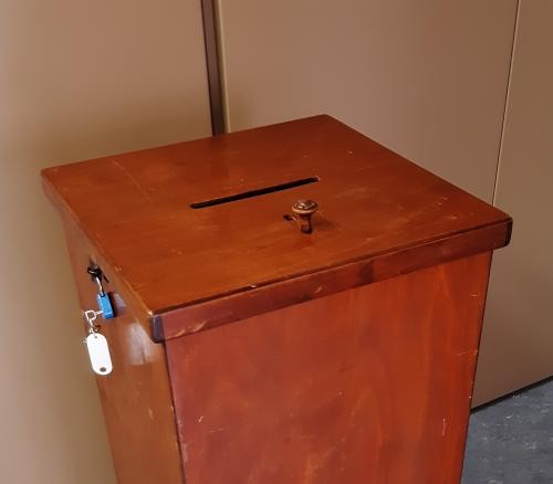 Es ist der obere Teil einer braunen, hölzernen Wahlurne zu sehen. Oben ist ein Einwurfschlitz mit einem Hebel. An der Seite hängt ein Marderschloss mit einem steckenden Schlüssel.