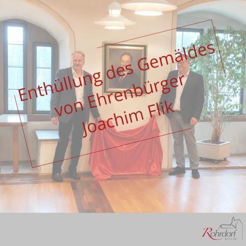 Im Rathaus der Gemeinde Rohrdorf stehen zwei Männer neben dem enthüllten Porträt von Ehrenbürger Joachim Flik. Unter dem Gemälde liegt ein rotes Tuch. Über dem Bild steht in roter Schrift: ‚Enthüllung des Gemäldes von Ehrenbürger Joachim Flik‘. Die Aufnahme zeigt die feierliche Präsentation des Porträts im Rathaus.