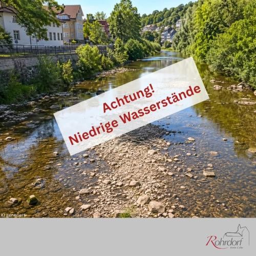 Ein Fluss mit sehr neidrigem Wasserstand zeigt zahlreiche freiliegende Kies- und Steinflächen. Entlang des Ufers befinden sich Bäume und Gebäude. Über dem Bild steht in einem weißen Hinweisfeld: "Achtung! Niedrige Wasserstände." Die Grafik weist auf die anhaltende niedrigen Wasserstände in Flüssen und Bächen hin